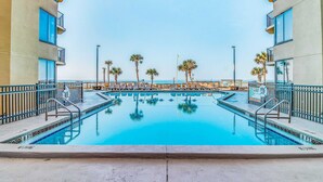 A swimming pool with clear blue water, surrounded by palm trees and a building with balconies. - Beautiful beachfront condo with private balcony and shared pool (Panama City Beach)