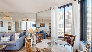 A modern living room with a glass dining table, a sofa, and a view of a cityscape through large windows. - Beautiful beachfront condo with private balcony and shared pool (Panama City Beach)