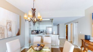 A modern kitchen with white cabinets, a central island, and a chandelier. There is a dining area with a glass table and white chairs. - Inviting Gulf-front condo In Perdido Key with waterfront pool/hot tub and oceanfront balcony (Perdido Key)