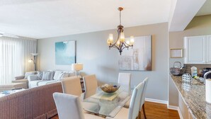 A modern living room with a glass dining table, white chairs, a sofa, and a chandelier. - Inviting Gulf-front condo In Perdido Key with waterfront pool/hot tub and oceanfront balcony (Perdido Key)