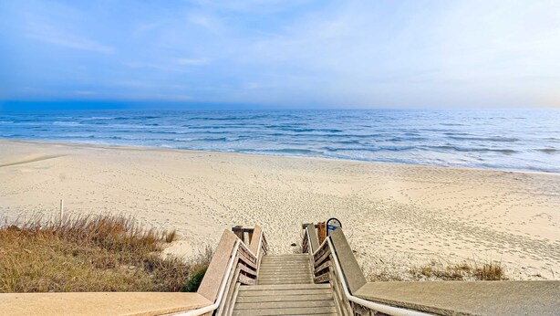 Eine Holztreppe führt zu einem Sandstrand, im Hintergrund das Meer.