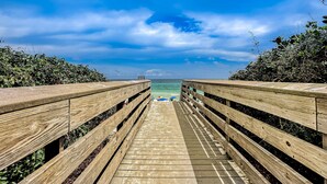 Ein Holzsteg führt zu einem Strand, im Hintergrund ist ein klarer Himmel und ein weiter Ozean zu sehen.