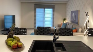 A modern living room with a kitchen area, featuring a dining table, chairs, a sofa, a TV, and a fruit bowl. - Sky Suites Mirage (Accra)