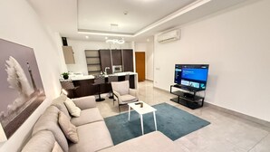 A modern living room with a sofa, a coffee table, and a TV. There is a kitchen area in the background with cabinets and a microwave. - Sky Suites Mirage (Accra)