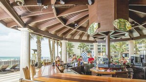 Ein Außenbereich mit Bar, Holzsitzmöbeln, einer Theke mit Barkeeper und Blick auf den Strand.
