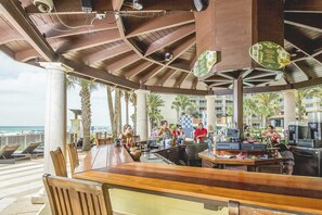 An outdoor bar area with wooden furniture, a counter with a bartender, and a view of the beach. - Luxury penthouse suite with Gulf-view wraparound balcony, pools, hot tubs & AC (Panama City Beach)