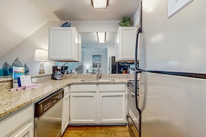 A modern kitchen with white cabinetry, a granite countertop, stainless steel appliances, and a view into the living area. - Gulf-front third-floor condo with WiFi, pool, beach boardwalk & central AC (Destin)