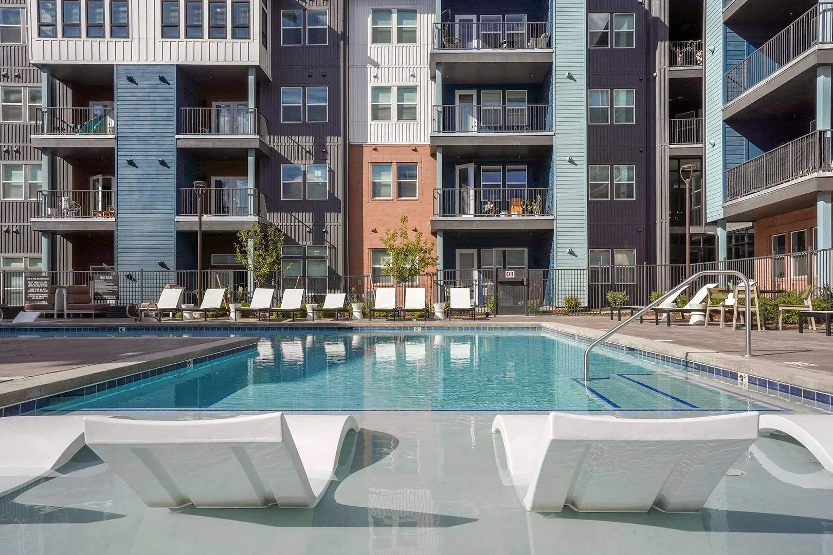 A modern apartment building with a swimming pool, multiple balconies, and lounge chairs.