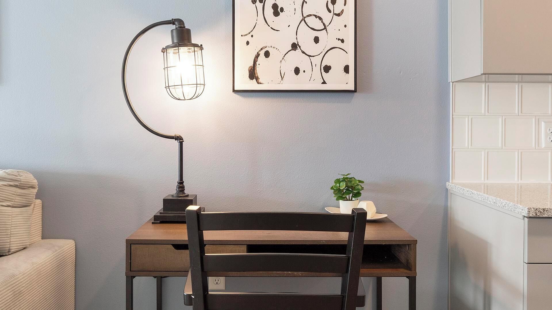 A small wooden table with a black chair, a desk lamp, a potted plant, and a framed abstract art piece on the wall.