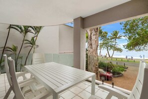 Ein Balkon mit weißem Tisch und Stühlen, der Blick auf einen Strand mit Palmen und einem klaren Himmel bietet.