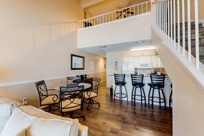 A spacious living area with a dining table and chairs, a kitchen in the background, and a staircase leading to an upper level. - Oceanfront condo with AC, WiFi, pool & seasonal beach service (Destin)