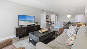 Ein Wohnzimmer mit Fernseher, dunklem Holzschrank, beiger Couch und gemustertem Sessel, sowie einem Essbereich mit einer Pendelleuchte.