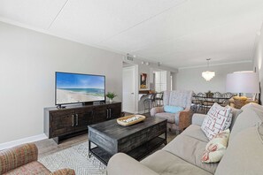 Ein Wohnzimmer mit Fernseher, dunklem Holzschrank, beiger Couch und gemustertem Sessel, sowie einem Essbereich mit einer Pendelleuchte.