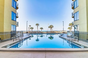 A swimming pool with a clear view of the ocean, surrounded by palm trees and a building with balconies. - Designer beachfront Gulf-view condo - beach access, pools, tennis, shuffleboard (Panama City Beach)