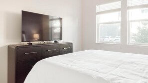 A hotel room with a bed, a dark wooden dresser with drawers, a flat-screen TV mounted above the dresser, and a window with a view of parked cars outside. - Landing | Spectacular 1BD, Pool, Gym (Colorado Springs)