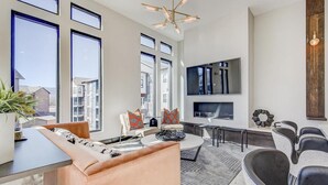 Una sala de estar amplia con ventanas grandes, una lámpara moderna, un televisor de pantalla plana y un rincón de estar con una mesa de centro.