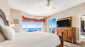 A large bed with a wooden headboard, a television on a wooden cabinet, and a view of the ocean through a window. - Spacious Gulf front condo w/shared hot tub, pools, gym - close to dining (Panama City Beach)