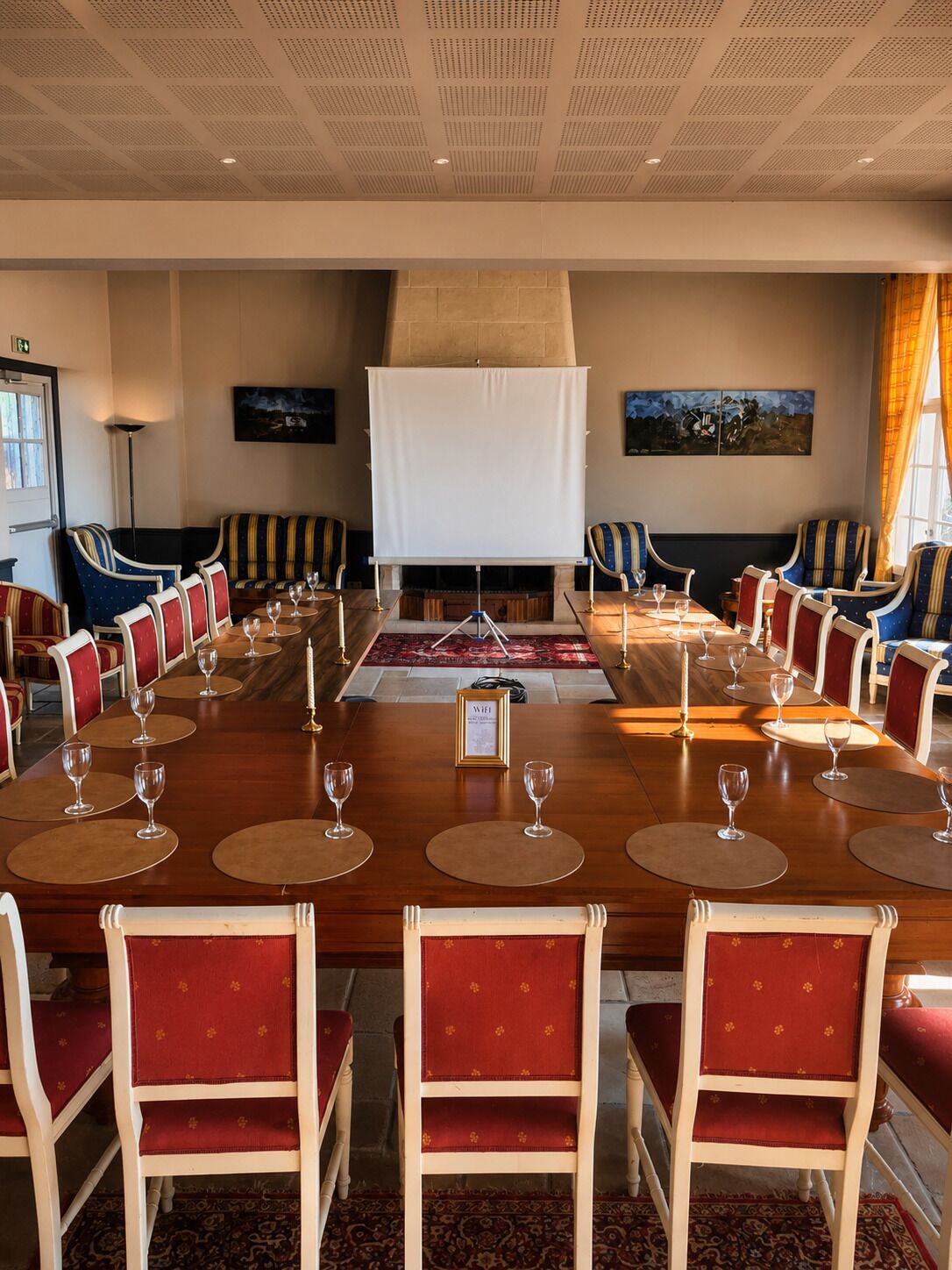 A conference room with a long table, red upholstered chairs, and a white projection screen.