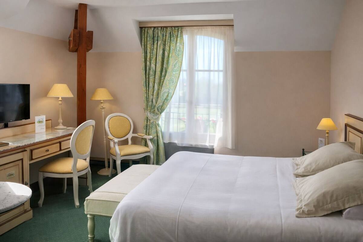 A hotel room with a bed, two chairs, a desk with a TV, and a window with curtains.