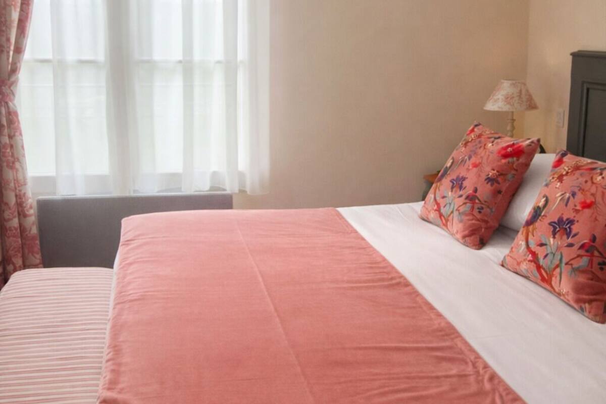 A bed with a pink bedspread and floral pillows, a bedside table with a lamp, and a window with sheer curtains.