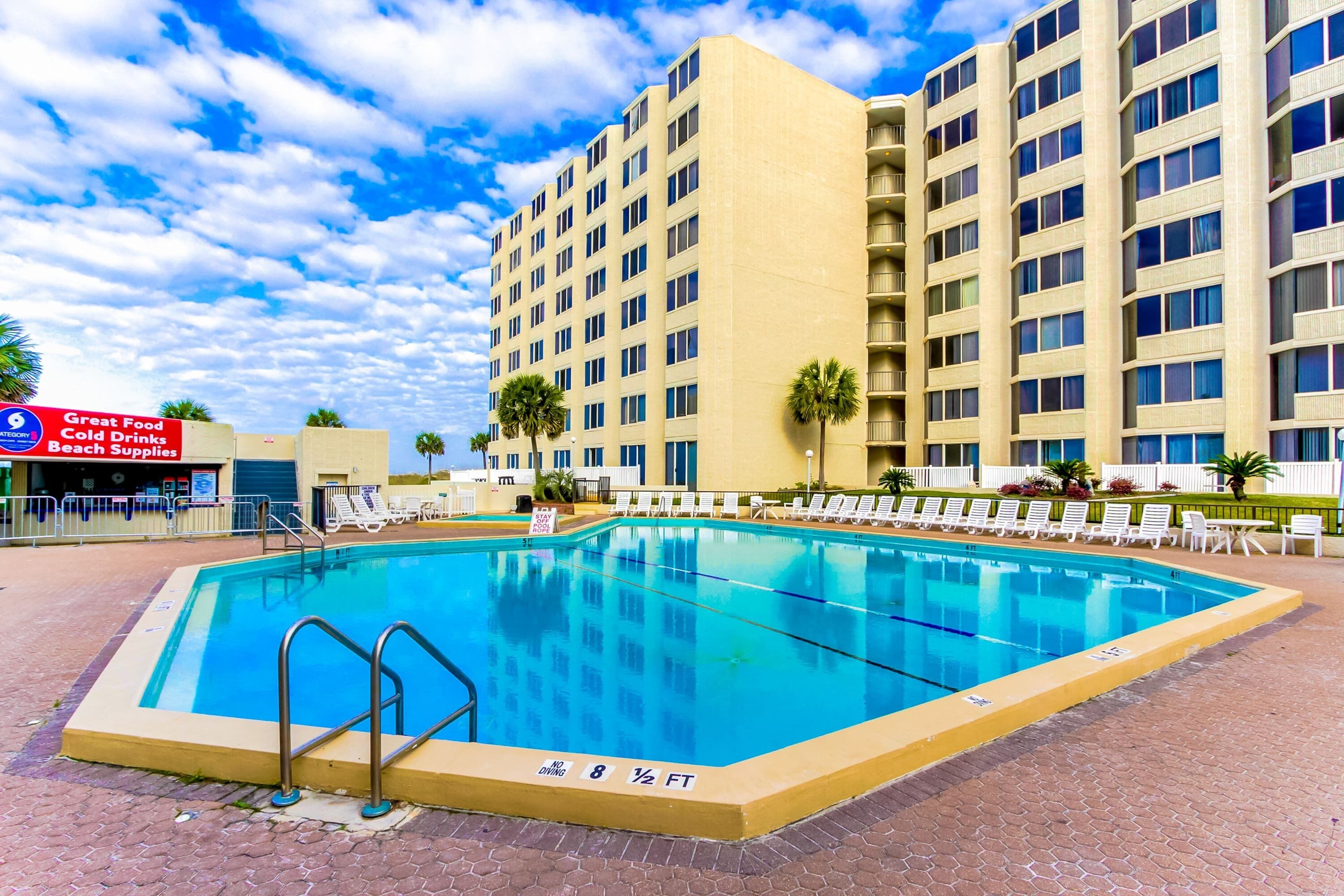A swimming pool with a no diving sign, surrounded by lounge chairs and palm trees, with a hotel building in the background.