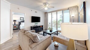 A modern living room with a sofa, coffee table, and a TV. There is a ceiling fan, a lamp, and a view of the outside through large windows. - Welcoming, Coastal Condo On Okaloosa Island Playground, 2 Hot Tubs (Fort Walton Beach)