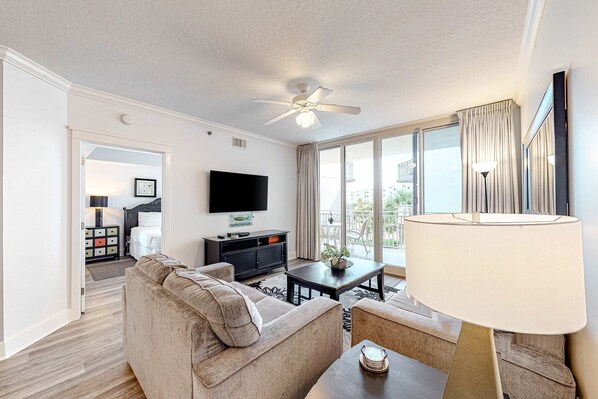 Un salon moderne avec un canapé, une table basse et un téléviseur. On y trouve un ventilateur de plafond, une lampe, et une vue sur l’extérieur grâce à de grandes fenêtres.