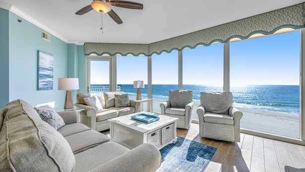 A spacious living room with a view of the ocean, featuring a ceiling fan, a large window, and a light-colored sofa set.