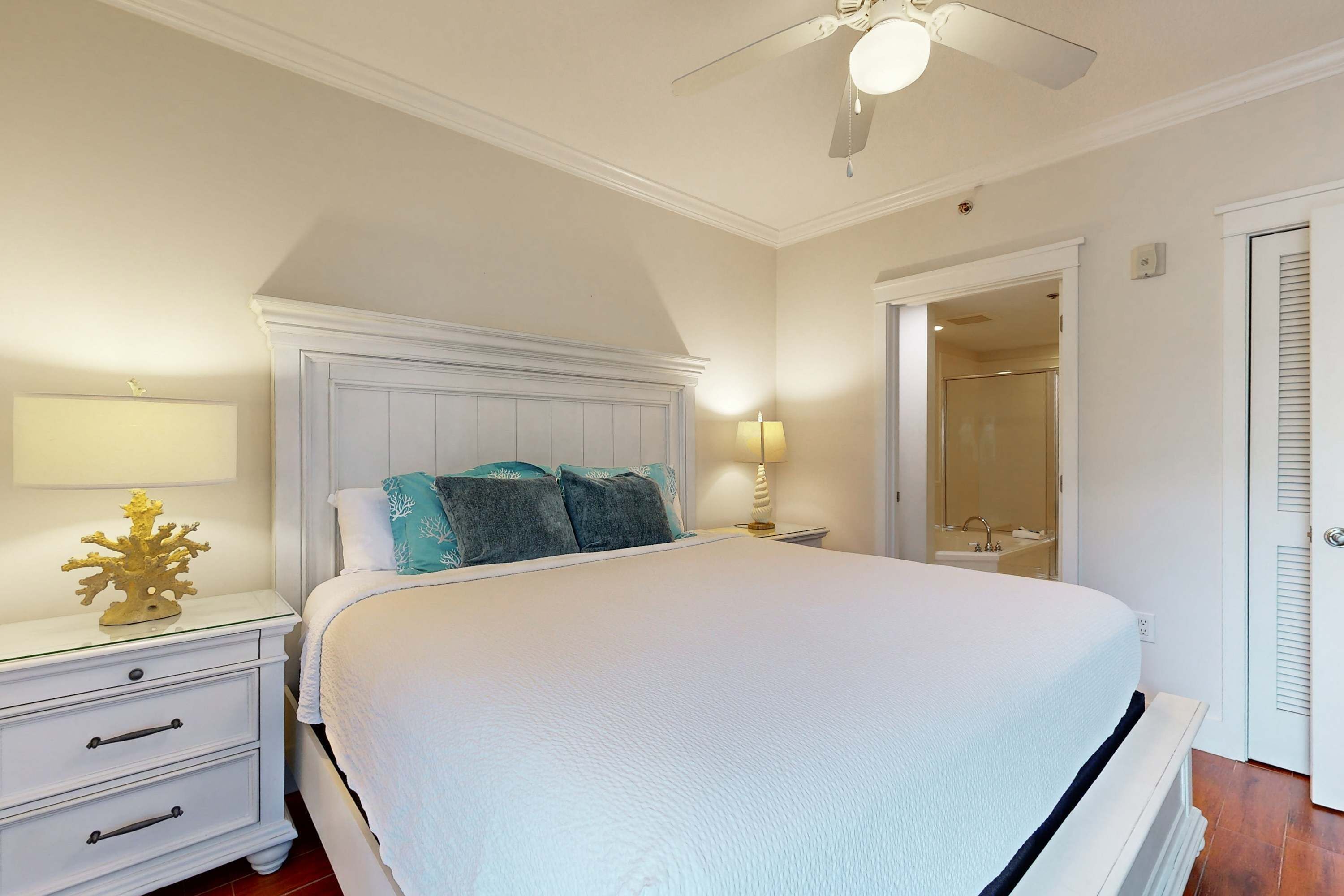 A bedroom with a large bed, a nightstand, a lamp, a ceiling fan, and a door leading to a bathroom.