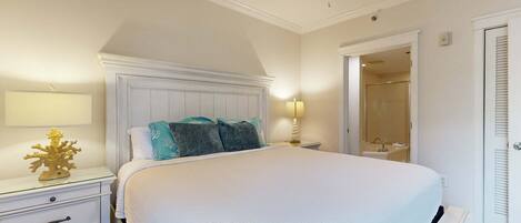 A bedroom with a large bed, a nightstand, a lamp, a ceiling fan, and a door leading to a bathroom.