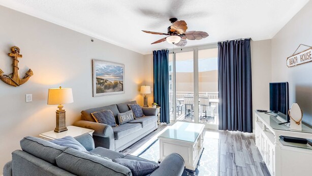 Una sala de estar moderna con un sofá, un sillón y una mesa de centro. Hay un ventilador de techo, una lámpara y un letrero que dice 'BEACH HOUSE'.