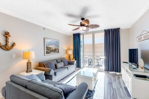 A modern living room with a sofa, armchair, and coffee table. There is a ceiling fan, a lamp, and a sign that reads "BEACH HOUSE". - Inviting, Coastal Condo On Okaloosa Island Waterfall, Lazy River (Fort Walton Beach)