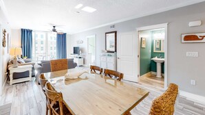 Un salón amplio con una mesa de comedor de madera, un sofá y un televisor. Se ve un baño con un lavamanos y un espejo a través de una puerta abierta.