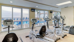 Une salle de sport équipée de machines de cardio et offrant une vue sur une piscine et des chaises longues.