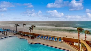 Un espace piscine doté de chaises longues et de palmiers, offrant une vue sur une plage et l’océan.