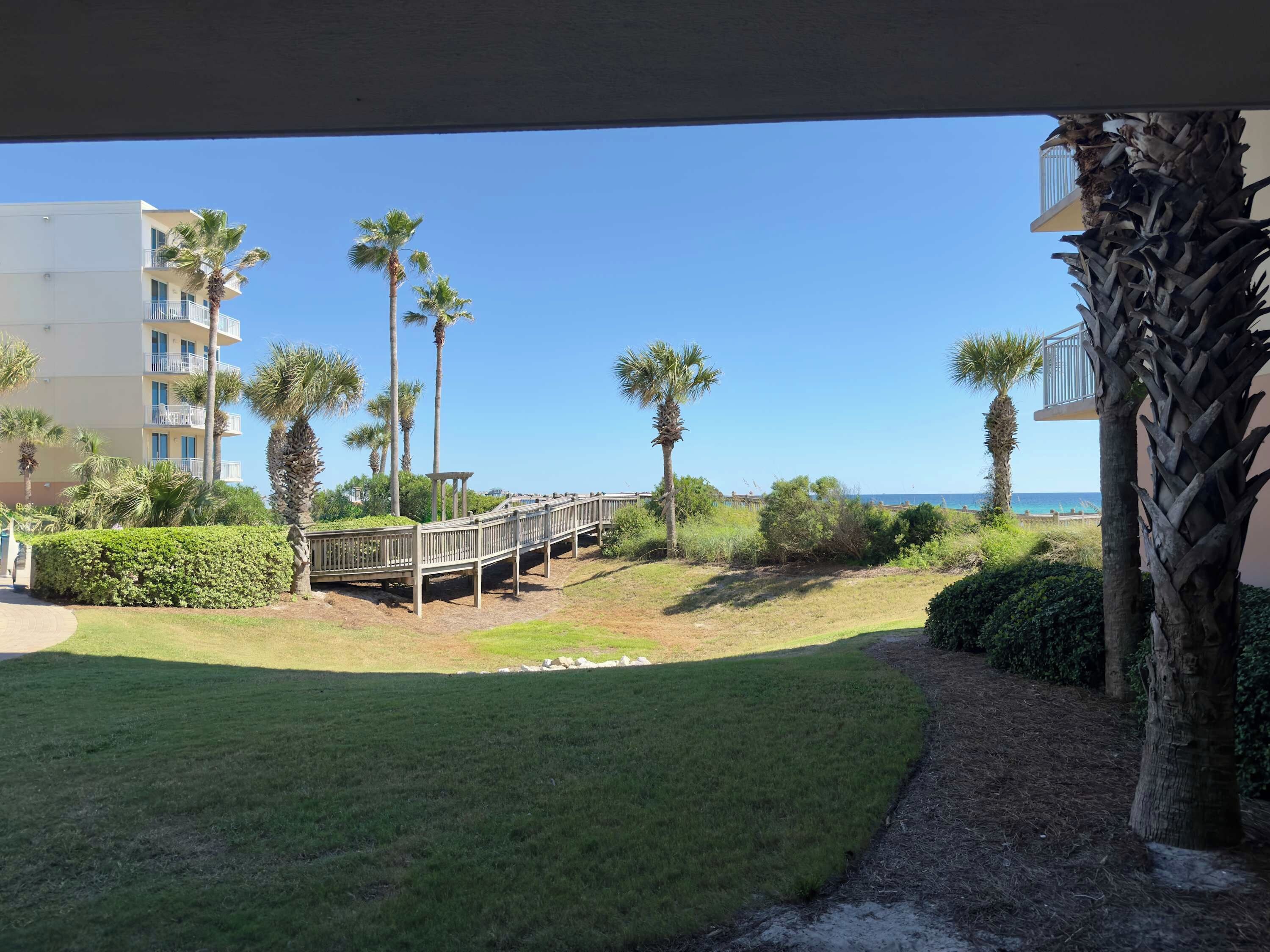 A view of a resort with a wooden deck, palm trees, and a building in the background.