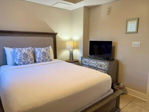 A neatly made bed with a headboard, a bedside lamp, a television on a stand, and a small trash bin. - Sunny Waterscape ground-floor condo with pools, hot tubs, & stunning Gulf views (Fort Walton Beach)