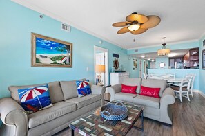 A living room with a ceiling fan, a dining area with white chairs, and a kitchen in the background. - Fifth Floor Condo with Gulf Views, Shared Pool and Hot Tub, and High-Speed WiFi (Fort Walton Beach)