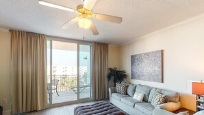 Una sala de estar con sofá, reposapiés, ventilador de techo y un ventanal con cortinas.