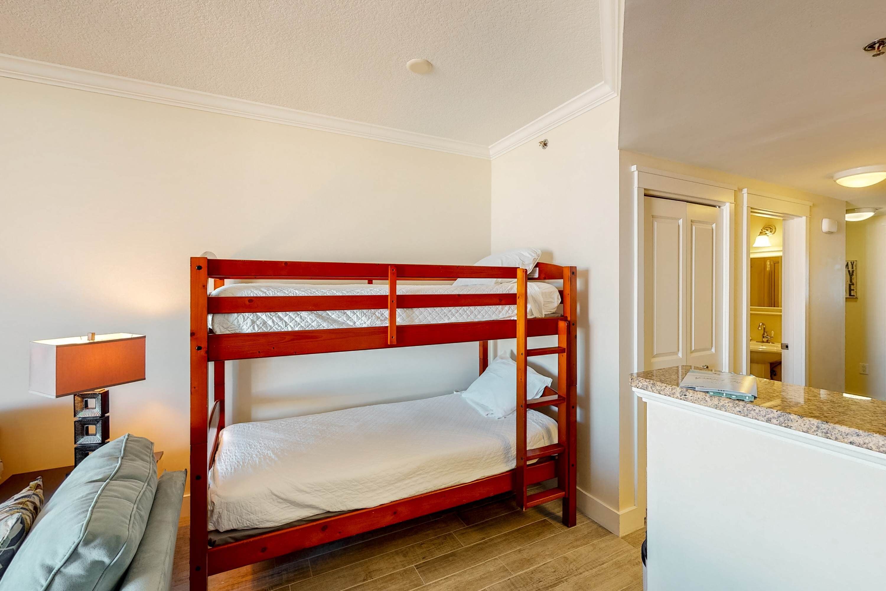 Een stapelbed met houten frame, een nachtkastje met lamp, en een badkamer zichtbaar door een geopende deur.