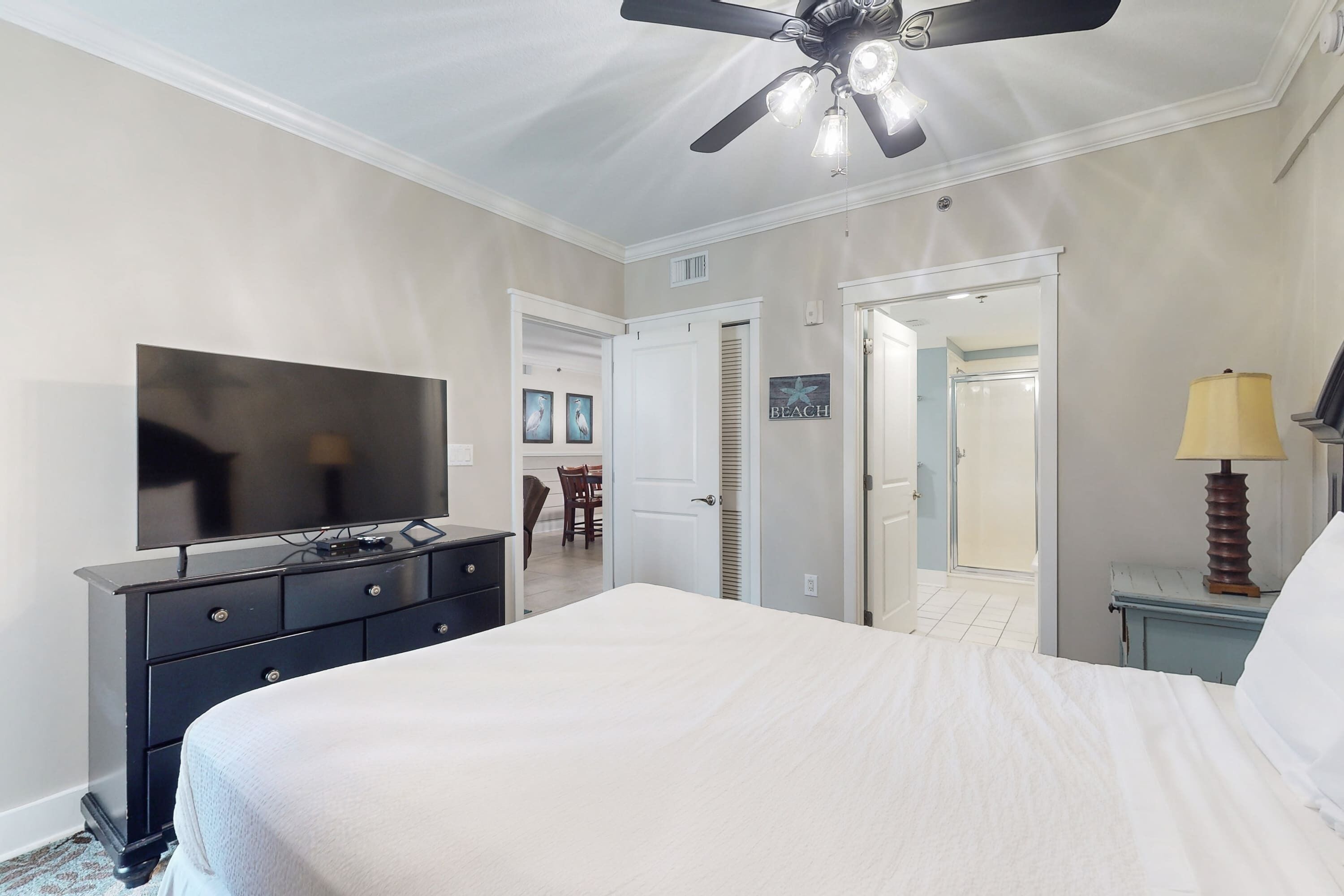 Une chambre à coucher comprenant un lit, un téléviseur, un ventilateur de plafond, une lampe et une porte donnant sur une salle de bain.