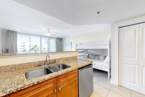 A modern kitchen with granite countertops, a double sink, and a dishwasher. There is a bunk bed with a white headboard in the background. - 3rd Floor Spacious, Beachy Condo At Waterscape Waterfall, Lazy River (Fort Walton Beach)