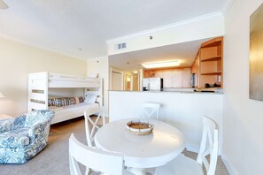 Un espace de vie compact comprenant un coin cuisine, un coin repas avec une table ronde et des chaises, un canapé blanc et un lit superposé.