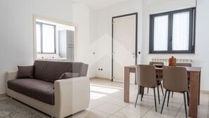 A modern living room with a sofa, a dining table with chairs, a large window with curtains, and a door.
