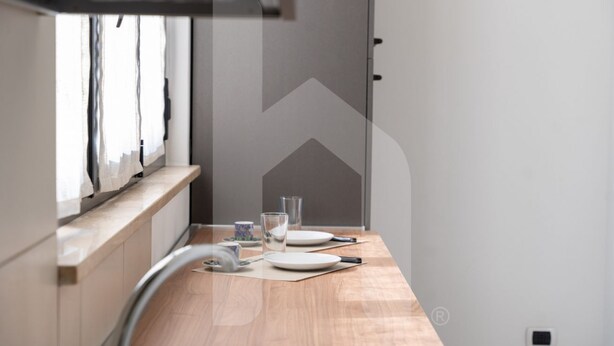 A modern kitchen with a wooden countertop, a window with sheer curtains, and a dining area with a table set for two.