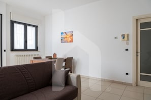 A modern living room with a sofa, a dining table, and chairs. There is a painting on the wall and a door with a glass panel. - Homiday - Maximum's Landing (Roseto degli Abruzzi)