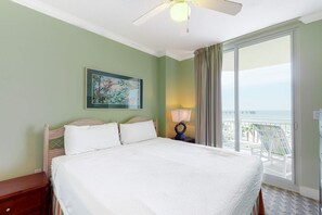 Ein Hotelzimmer mit Bett, Nachttisch, Lampe, einem Gemälde und Meerblick durch ein großes Fenster.