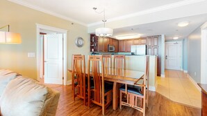 A spacious living room with a wooden dining table and chairs, a sofa, and a kitchen area with wooden cabinets and appliances. - 5th Floor Gulf-Front Condo w Free WiFi, Shared Hot Tub, Outdoor Pool, & More (Fort Walton Beach)