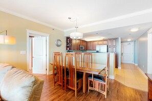 A spacious living room with a wooden dining table and chairs, a sofa, and a kitchen area with wooden cabinets and appliances. - 5th Floor Gulf-Front Condo w Free WiFi, Shared Hot Tub, Outdoor Pool, & More (Fort Walton Beach)