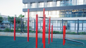 Ein Outdoor-Fitnessbereich mit roter Trainingsausstattung, darunter Klimmgeräte und eine Bank, vor einem modernen Gebäude mit großen Fenstern.